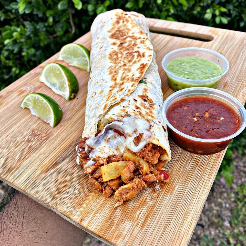 El Rucon's large burrito with melted cheese, limes, and two dipping sauces on a wooden board.