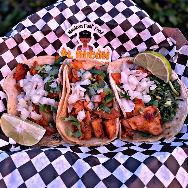Three El Rucon al pastor tacos with onions, cilantro, and lime wedges in checkered paper.