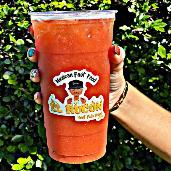 Hand holding a large El Rucon Mexican Fast Food drink with a green leafy background.