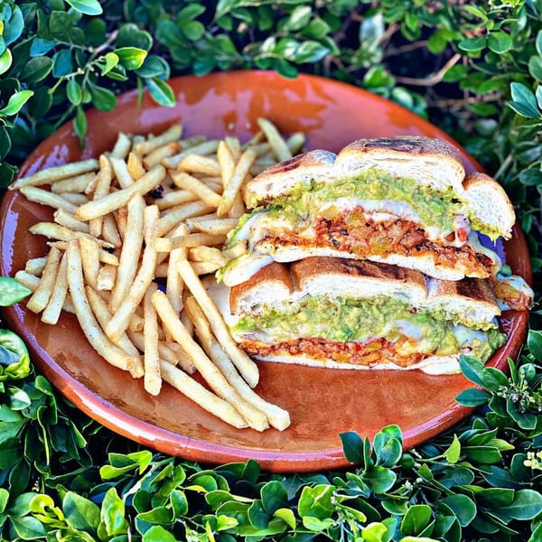 A Mexican torta sandwich with guacamole and melted cheese, served with fries on a rustic plate.