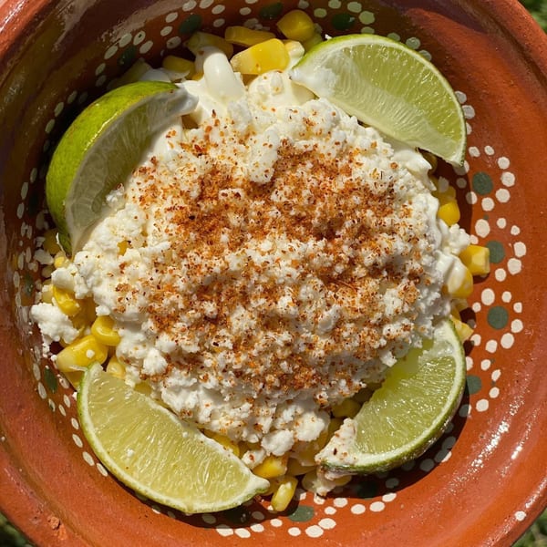 Mexican esquites in a terracotta bowl, topped with cotija cheese, chili powder, and lime wedges.